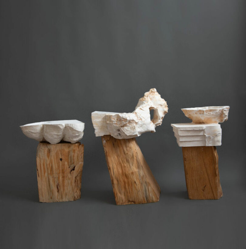 Tres esculturas abstractas de Dana Harel hechas de bloques de madera en bruto y moldes de yeso blanco texturizado se exhiben sobre un fondo gris sólido.
