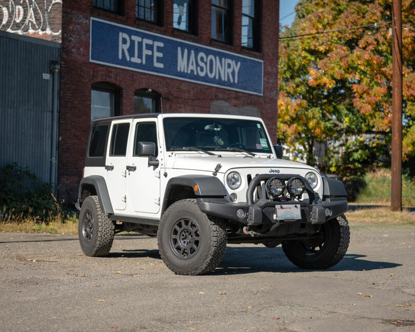 Un Jeep Wrangler blanco estacionado en un terreno pavimentado frente a un edificio de ladrillo con un "Albañilería popular" símbolo.
