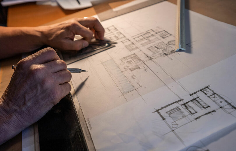 Un hombre dibuja dibujos arquitectónicos en papel de calco con una regla y un portaminas en un escritorio.