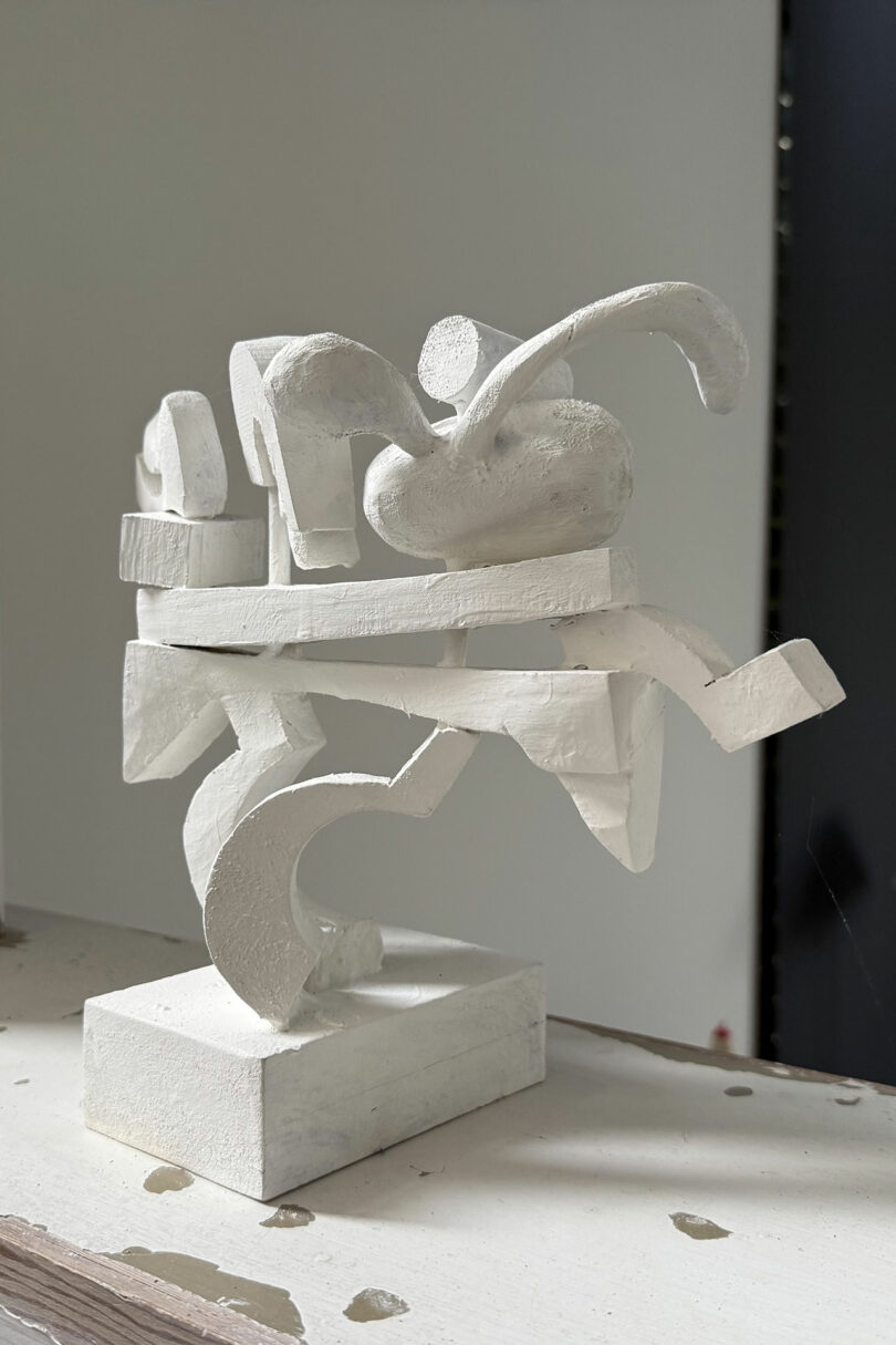 Escultura blanca abstracta con formas geométricas y curvas que se cruzan sobre una base blanca sobre un fondo liso.