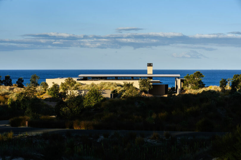 Una casa moderna con techo plano está ubicada entre la vegetación costera, con vistas al mar bajo un cielo azul y nubes blancas.