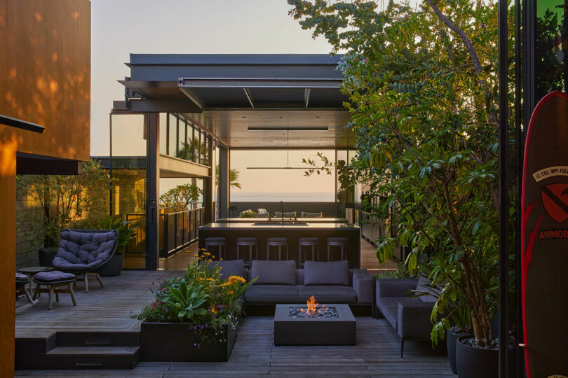 El contemporáneo patio al aire libre cuenta con muebles modernos, una fogata, exuberantes plantas y un área de cocina abierta frente al océano al atardecer.