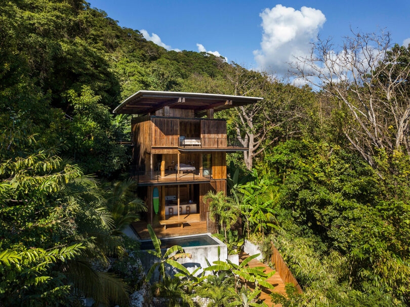 Moderna casa de madera de tres pisos con grandes ventanales y una pequeña piscina rodeada de un exuberante bosque tropical y colinas bajo un cielo azul.