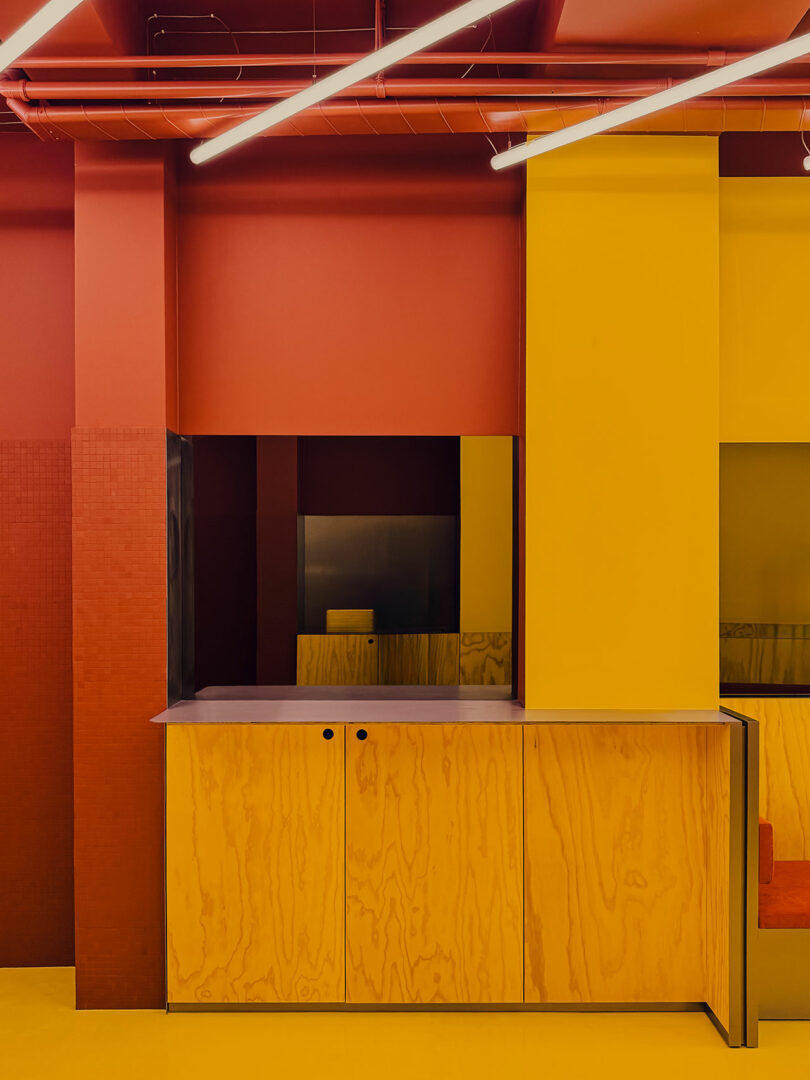 Interior minimalista con paredes y armarios amarillos, conductos de techo rojos y luces de techo lineales con textura de madera natural y elementos de diseño geométrico.