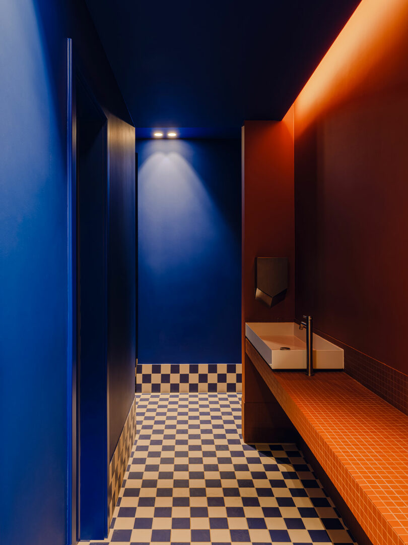 El moderno baño cuenta con paredes azules y naranjas, un piso a cuadros azules y blancos, un lavabo rectangular y un espejo de pared con una iluminación cálida.