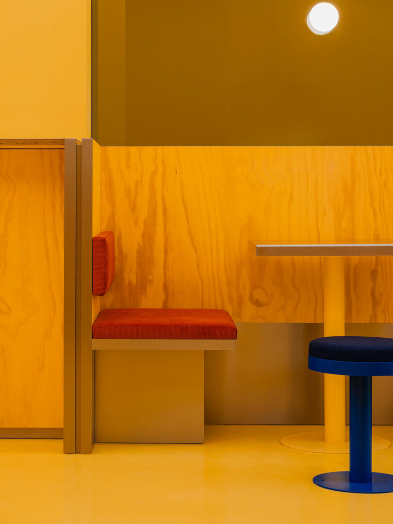 Interior minimalista de cafetería con paredes amarillas, una cabina de madera con cojines naranjas, una mesa de metal y taburetes azules bajo una lámpara de pared redonda.