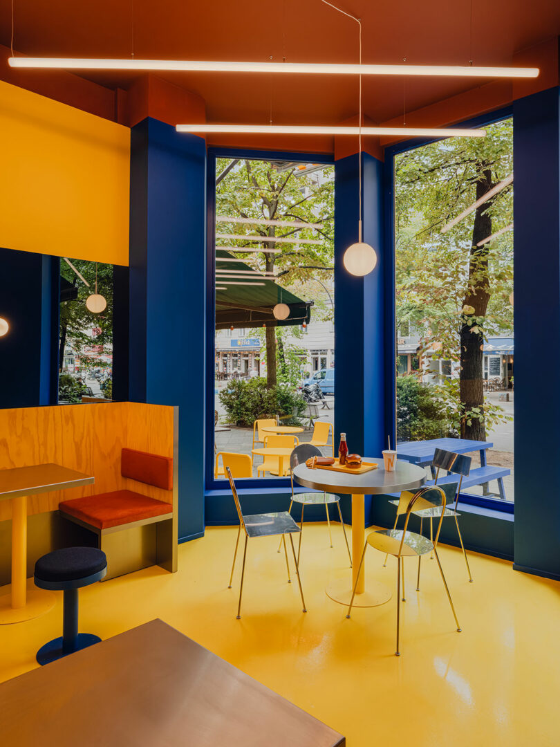 Interior colorido y moderno de una cafetería con suelo amarillo, paredes azules y grandes ventanales con árboles en el exterior. Se dispusieron varias mesas y sillas, y se colocaron bebidas y vajillas en las mesas redondas.