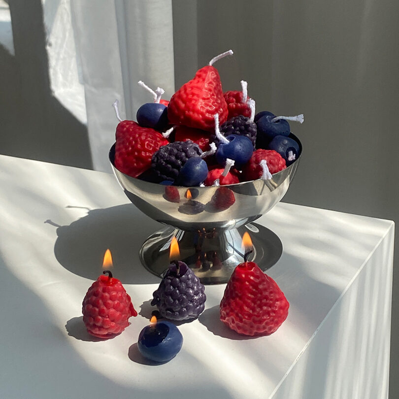 Un cuenco plateado lleno de velas con forma de frutas rojas y moradas se asienta sobre una superficie blanca; algunas velas se encienden y se colocan al frente, lo que lo convierte en una adición encantadora a cualquier guía de regalos de 2025.