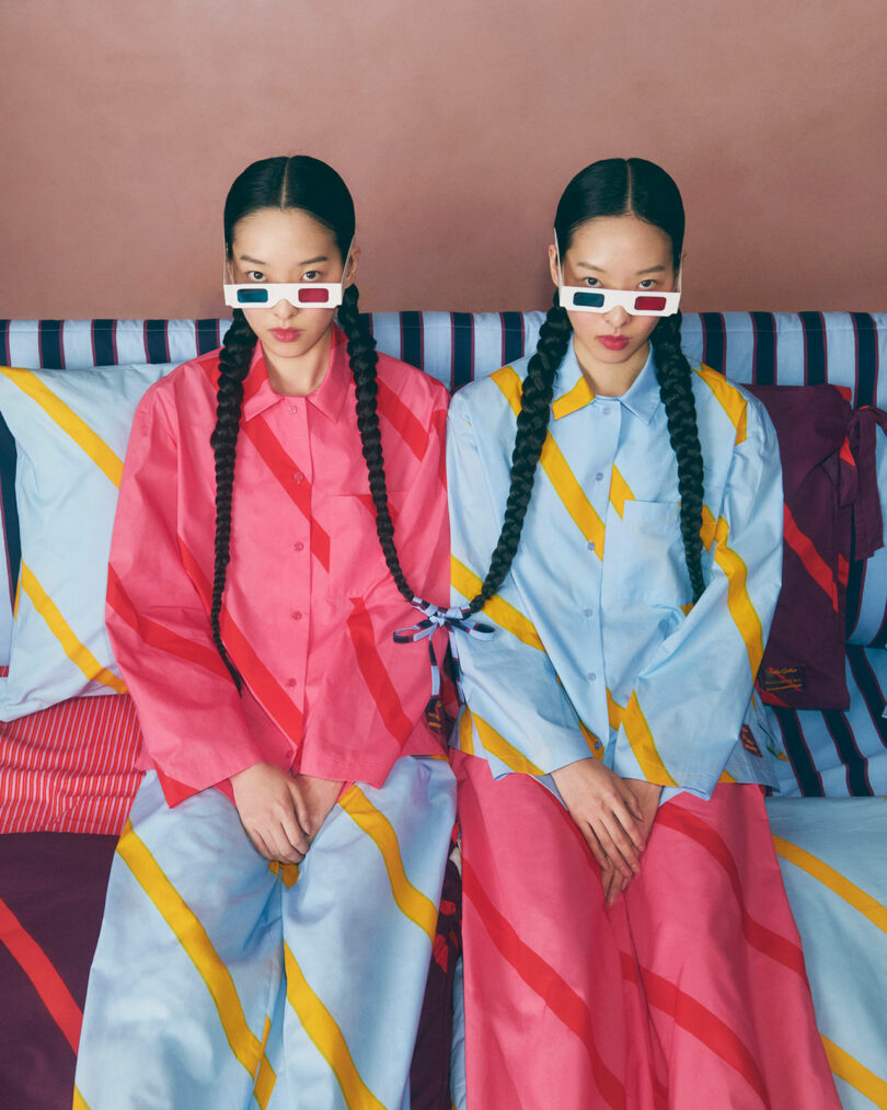 Dos mujeres trenzadas se sientan una al lado de la otra en una cama a rayas, vestidas con trajes estampados de colores brillantes y gafas 3D retro.