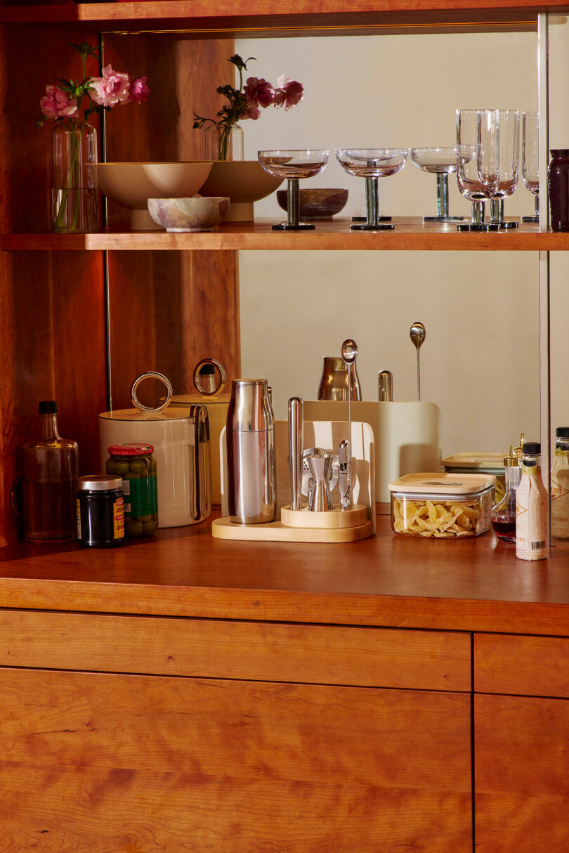 A wooden kitchen counter with jars, bottles, a utensil holder, and a tray; upper shelves hold bowls, glasses, and vases with pink flowers—perfect inspiration for your next Gift Guide—reflected beautifully in a mirror.