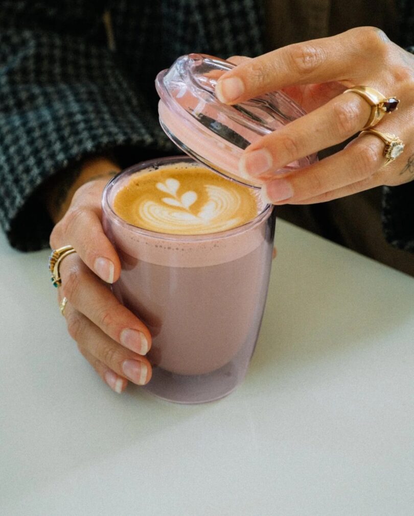 Una persona abre la tapa de un vaso rosa claro de doble pared que contiene una taza de café con leche con arte latte en forma de corazón