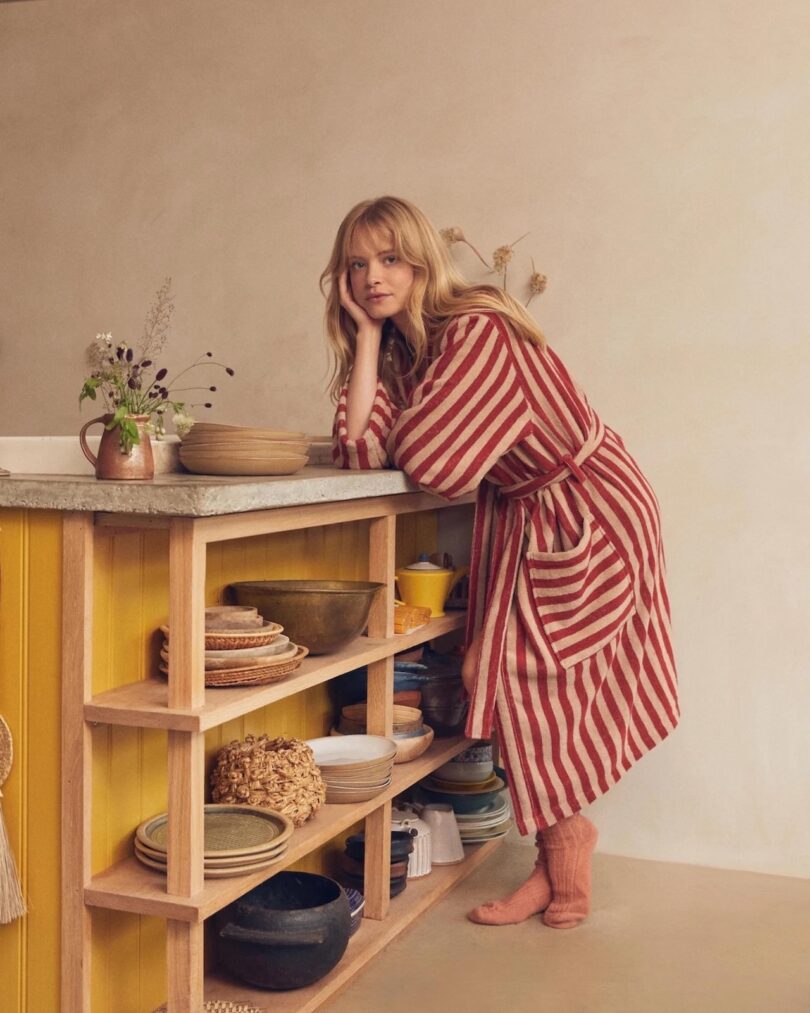 Una mujer de cabello rubio con una bata a rayas rojo-beige se apoya en la encimera de la cocina cubierta con platos, tazones y un jarrón de flores.