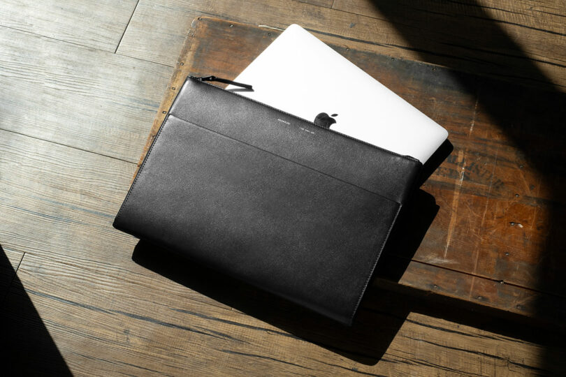 Funda cerrada de cuero negro para portátil que cubre parcialmente el MacBook plateado abierto sobre una superficie de madera a la luz del sol.