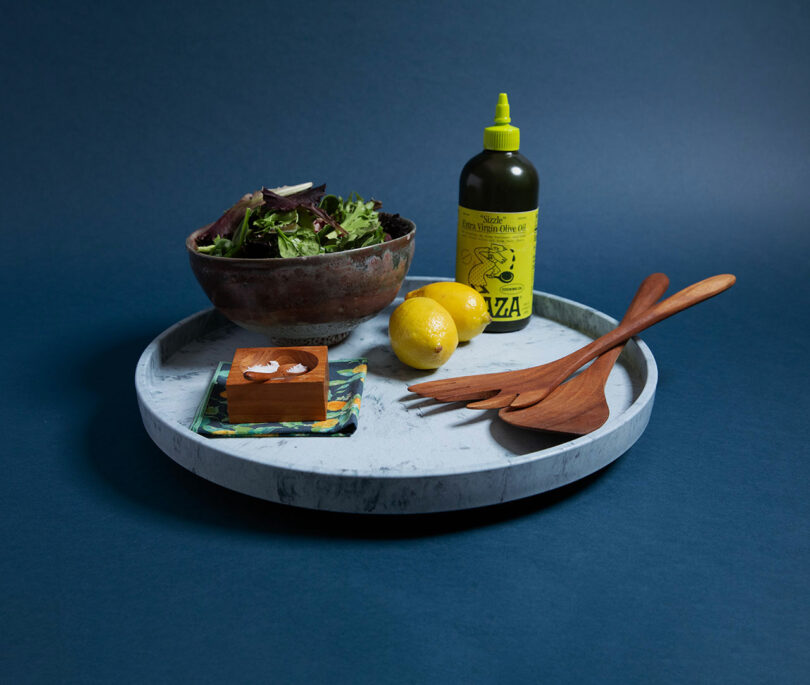A bowl of mixed greens, two lemons, a bottle of vinaigrette, a small wooden box, and wooden salad servers on a round marble tray against a blue background.