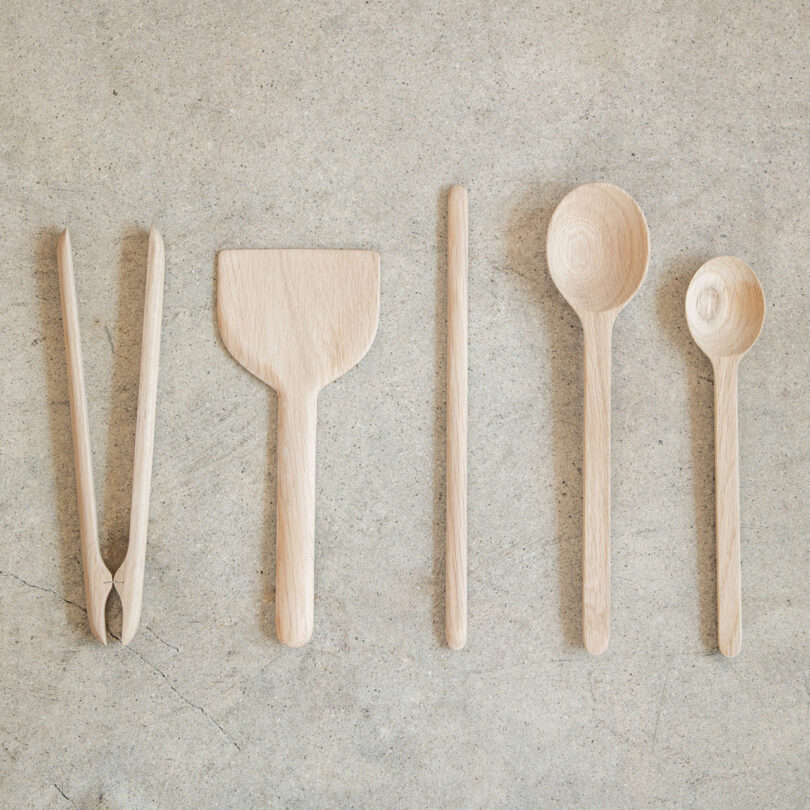 Cinco piezas de utensilios de cocina de madera, que incluyen pinzas, espátulas, palitos, cucharas grandes y cucharas pequeñas, dispuestas en fila sobre una superficie de textura clara, perfectas como regalo para un chef.