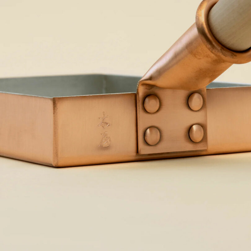 Primer plano de una olla cuadrada de cobre con caracteres japoneses grabados en el lateral y mango remachado adjunto, lo que la convierte en un complemento único para cualquier cocina e ideal como regalo para chefs.