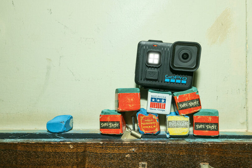 A GoPro camera, showcasing modern technology, sits atop several worn chalk boxes and pieces of blue chalk on a wooden surface, set against a plain light-colored wall.