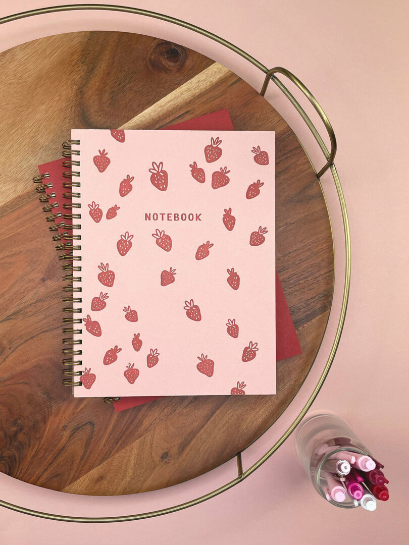 A pink spiral notebook with strawberry illustrations and "NOTEBOOK" on the cover sits on a wooden tray next to a glass of pink and red pens—perfect for your 2025 Gift Guide.