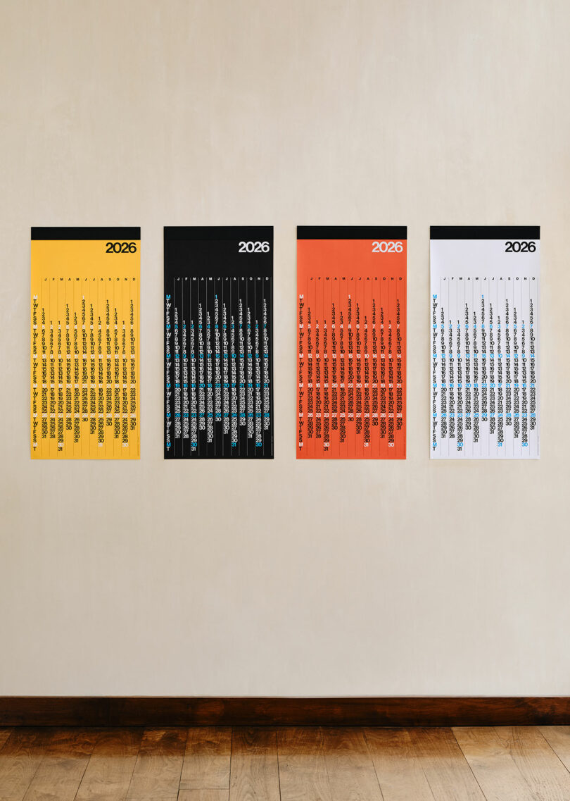 Four minimalist 2026 wall calendars in yellow, black, orange, and white are displayed side by side on a light-colored wall above a wooden floor.