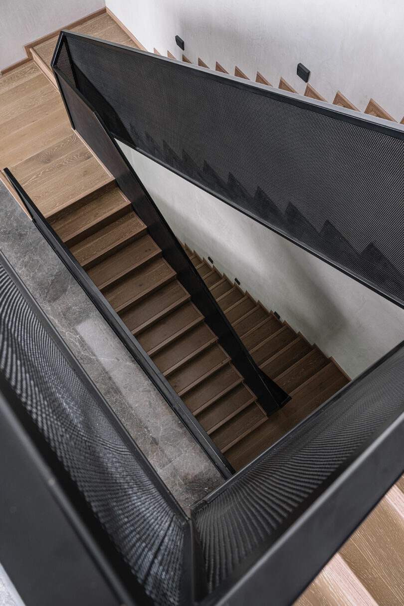 A modern wooden staircase with black metal mesh railings viewed from above, showing multiple flights and landings.