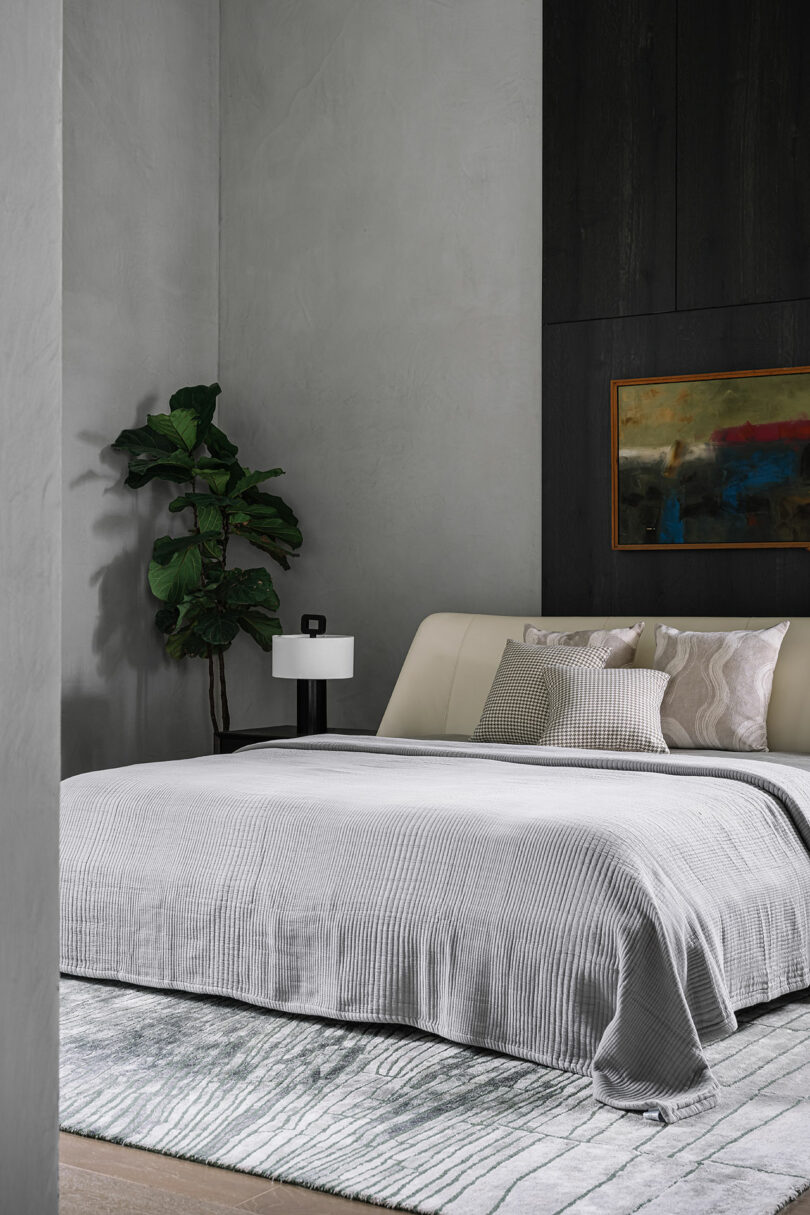 A modern bedroom with a low bed, neutral bedding, a potted plant, a white lamp on a black nightstand, and an abstract painting on the wall.
