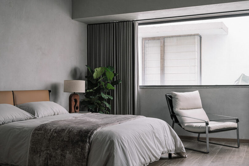 A minimalist bedroom with a neatly made bed, a side table with a lamp, a potted plant, an armchair, and a large window letting in natural light.