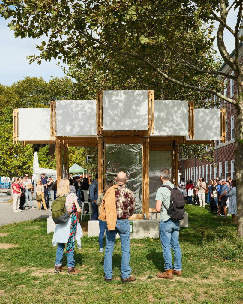 Un grupo de personas se reúne al aire libre, rodeado de árboles y un moderno pabellón de madera con grandes paneles de vidrio y elementos rectangulares blancos en el césped cerca del edificio.