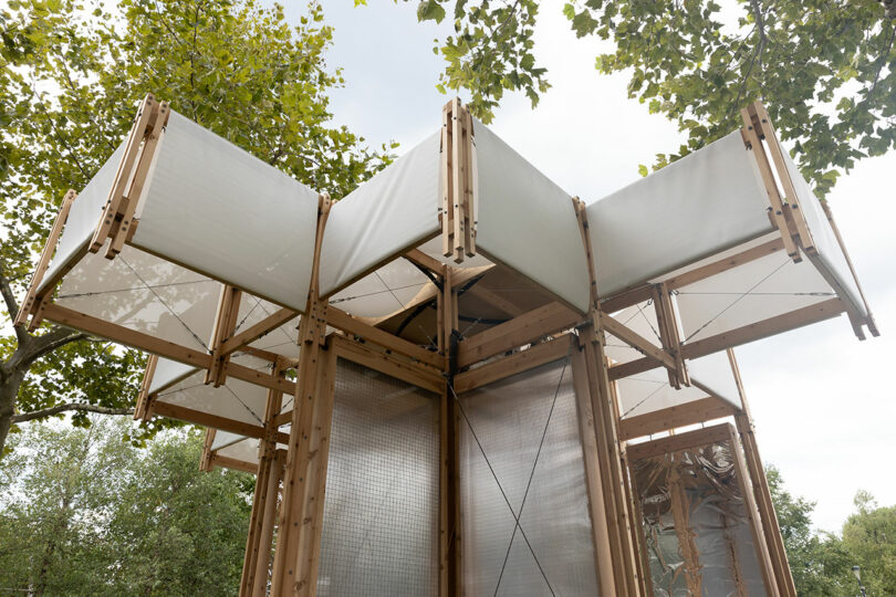 Una estructura de pabellón de madera con grandes paneles de tela blanca se extiende hacia afuera, rodeada de árboles verdes y contra un cielo nublado.