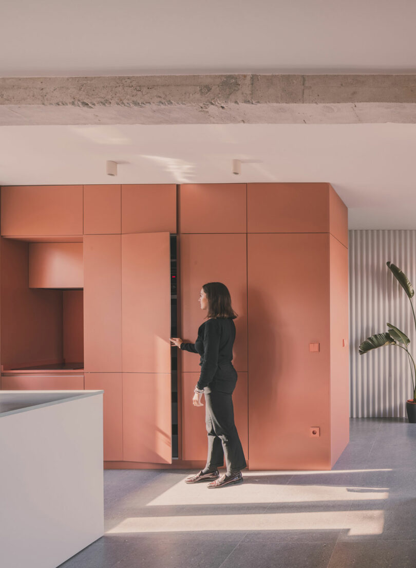 Un hombre vestido de negro abre un mueble alto en una cocina moderna con muebles de color naranja mate y diseño minimalista.