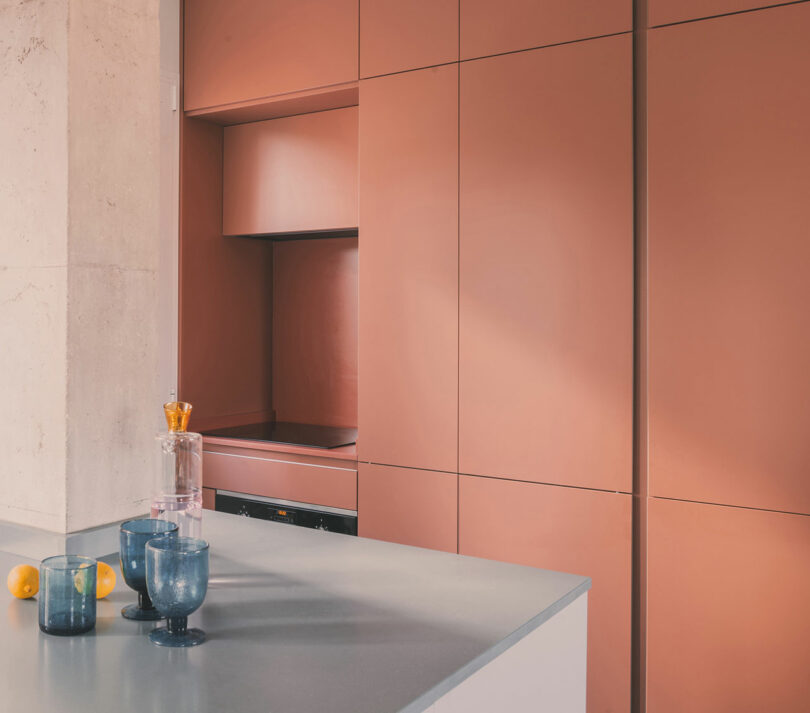 Cocina moderna con armarios de color rosa mate, encimeras grises, tres vasos azules, una jarra de cristal con tapón naranja y dos limones sobre la encimera.