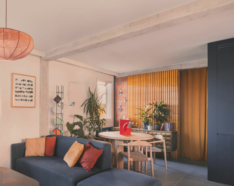 Moderna sala de estar y comedor con sofá seccional azul, cojines coloridos, mesa de comedor redonda, plantas de interior y grandes cortinas amarillas.