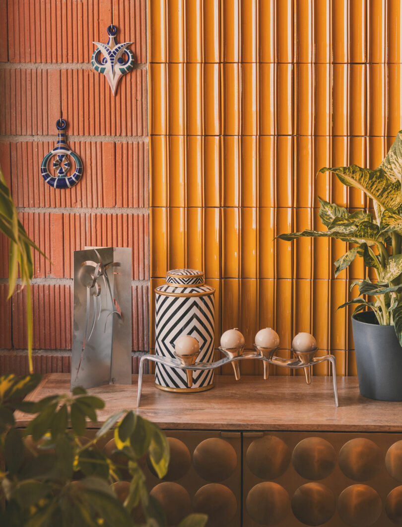 Mesa decorativa con un tarro geométrico, cuatro portahuevos, plantas en macetas y arte mural sobre azulejos amarillos y fondo de ladrillo rojo.