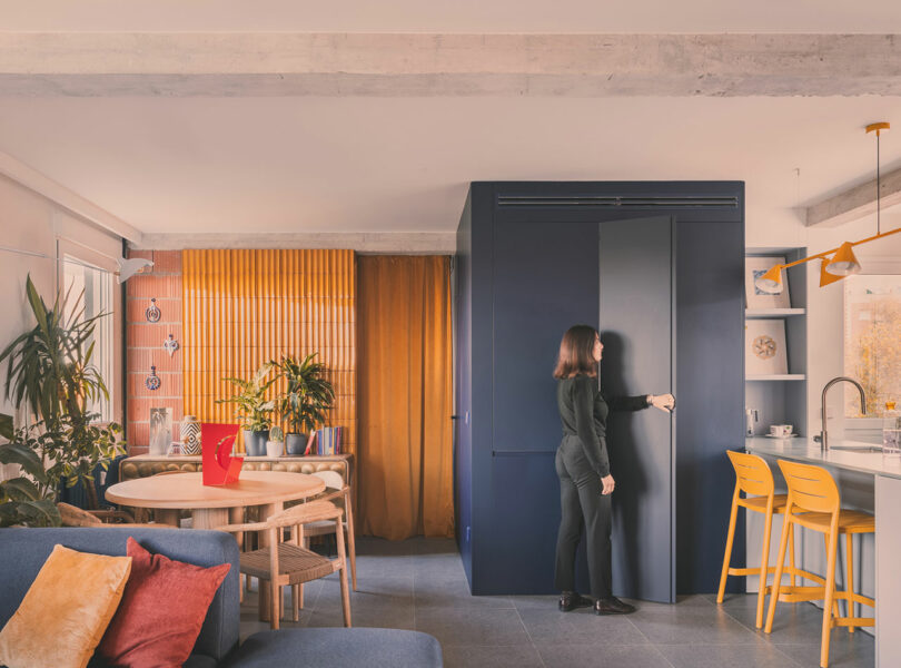 Una mujer se encuentra cerca de un gabinete azul oscuro en un apartamento moderno y colorido con tonos amarillos y naranjas, plantas y muebles modernos.
