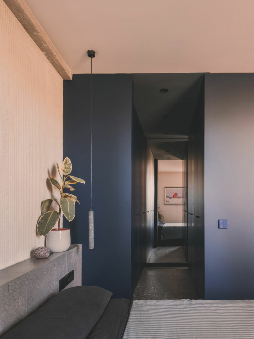 El moderno dormitorio cuenta con plantas en macetas sobre repisas de concreto, paredes de color azul oscuro, candelabros minimalistas y obras de arte enmarcadas visibles a través del estrecho pasillo.