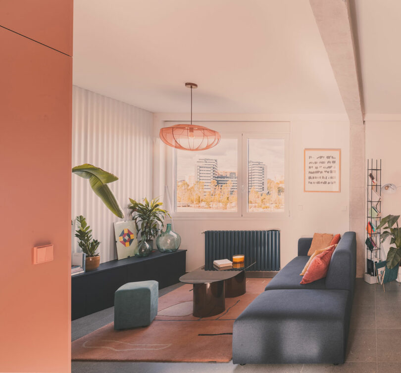 Salón moderno con sofá azul, mesa baja, plantas, alfombra geométrica y un gran ventanal con vistas a los edificios de la ciudad.