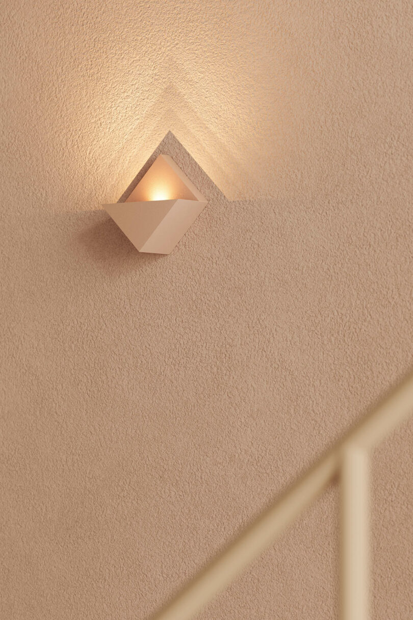 A modern wall sconce emits warm light, creating geometric shadows on a textured beige wall above a staircase railing.