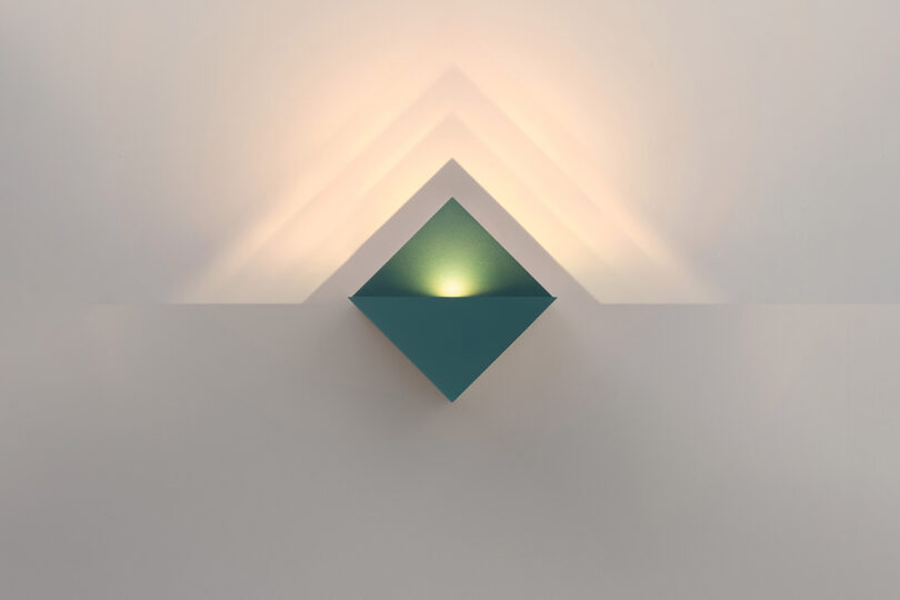 A teal, diamond-shaped wall sconce emits soft, upward and downward light, casting symmetrical shadows on a plain white wall.