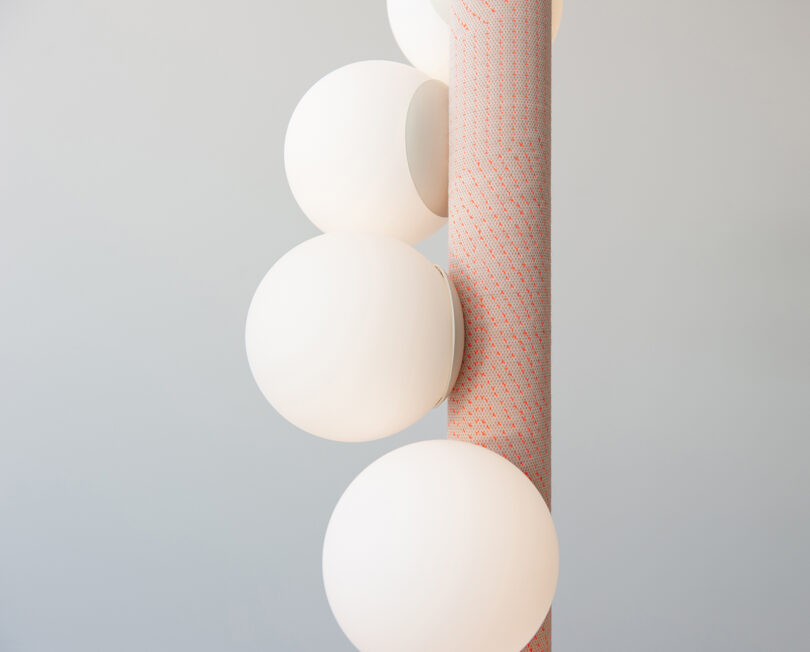 Una varilla vertical con una superficie texturizada sostiene cuatro luminarias esféricas blancas redondas montadas en un lado sobre un fondo neutro.