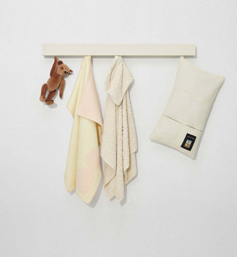 A plush toy, two patterned blankets, and a cream-colored pouch hang on a simple white wall rack.