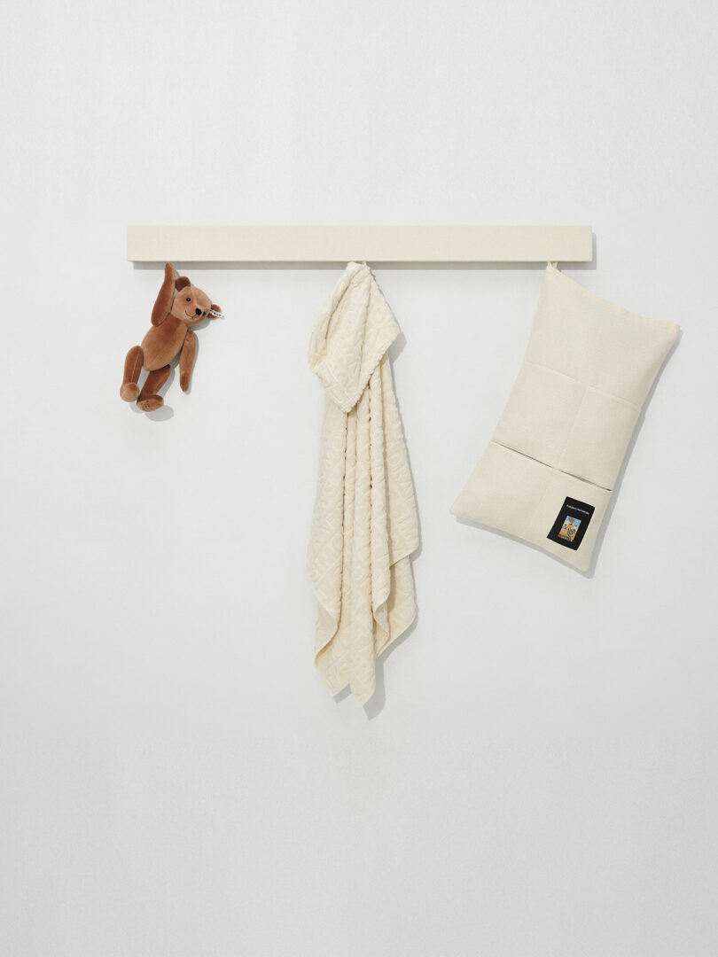 A brown plush rabbit, a cream-colored quilt, and a cream pillow with a label hang from a minimalist white wall rack.