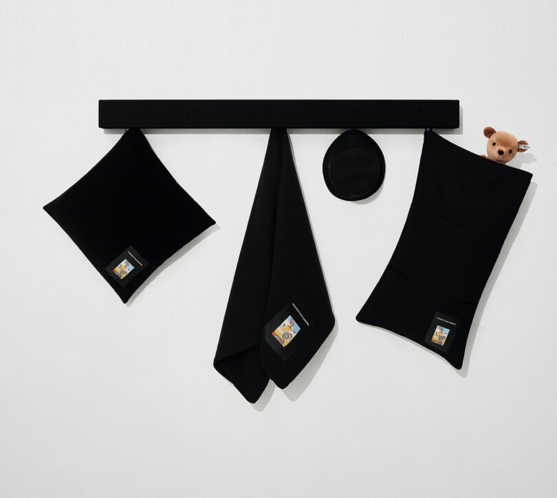 A black wall rack holds three black accessories with small labels; a small teddy bear peeks out from the rightmost pocket against a plain white background.