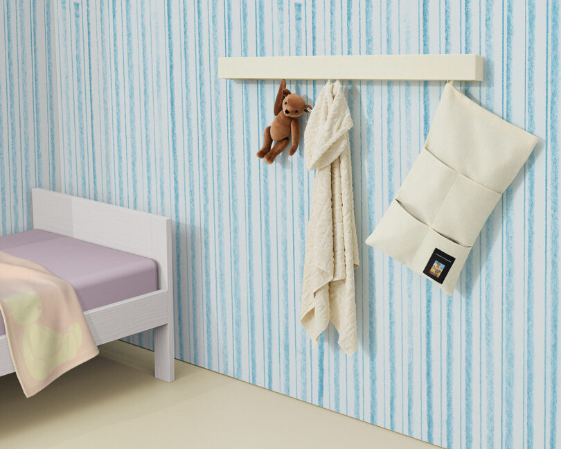 Junto a una pared con rayas azules hay una cama cuidadosamente hecha, con un estante blanco que sostiene un osito de peluche, dos trozos de tela colgantes y una bolsa blanca.