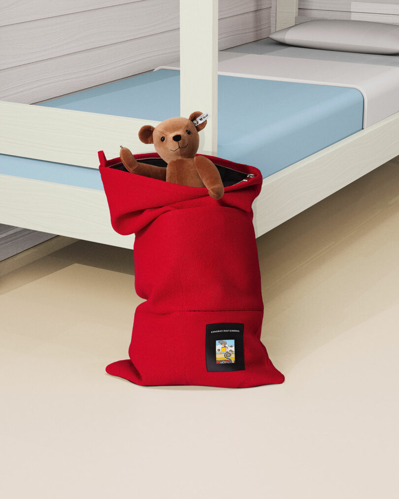 A brown teddy bear sits inside a red zippered bag placed on the floor next to a white and blue bunk bed.