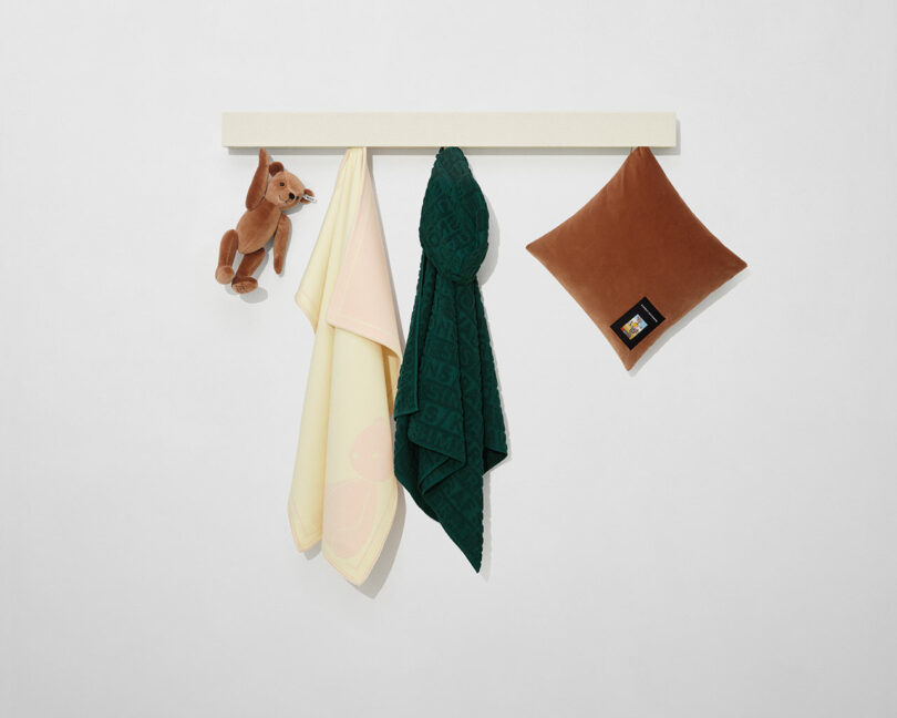 A wall-mounted white rack with a brown plush dog, a yellow cloth, a green textured towel, and a brown pillow hanging from it against a plain white background.