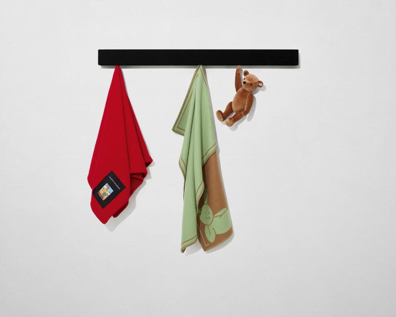A black wall-mounted bar holds a red blanket with a label, a green and brown blanket with a bunny design, and a brown plush toy hanging by one arm.