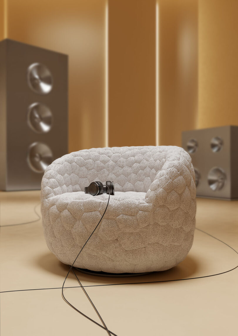 Un sillón redondo con textura descansa sobre un piso beige, con auriculares colocados en el asiento; Al fondo hay unos enormes altavoces plateados.