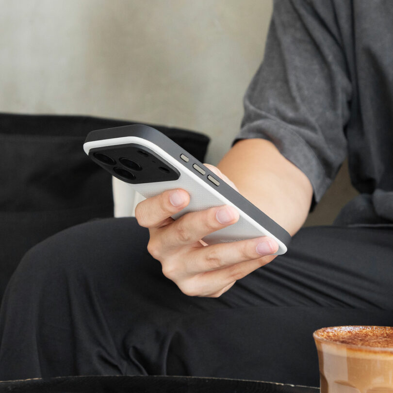 A person holding a smartphone with a two-tone case, seated at a table with a cup of coffee in the foreground.