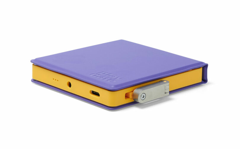 A compact, square electronic device with a purple cover, yellow sides, a metal latch, and ports for audio and USB connections. The word "playdate" is embossed on the cover.