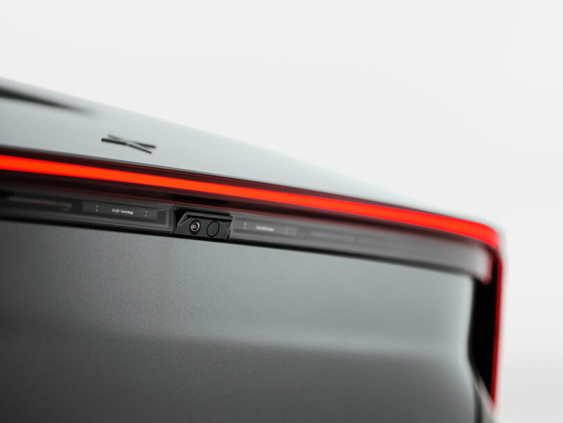 Primer plano de la cámara trasera del automóvil, integrada en las elegantes y modernas luces traseras con una tira LED roja continua sobre un fondo claro.