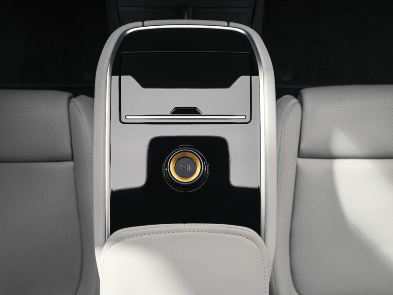 Un primer plano de la consola central trasera del automóvil, con molduras en negro brillante, botones de control circulares con un ícono de Reproducir/Pausa y asientos de cuero gris claro adyacentes.